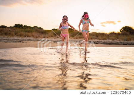 Joyful sisters splashing water during sunset beach adventure Joyful sisters splashing water during sunset beach adventure 128670460