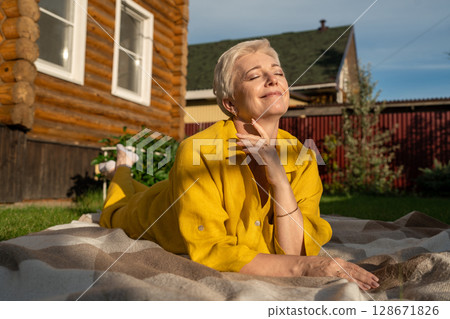 A serene scene of a mature woman dressed in yellow, leisurely relaxing on a blanket in her garden near house, enjoying sun A serene scene of a mature woman dressed in yellow, leisurely relaxing on a blanket in her garden near house, enjoying sun 128671826