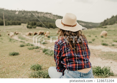 A woman in a plaid shirt and hat is sitting in a field with a herd of sheep 128672204