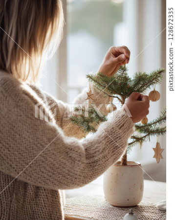 Faceless woman in cozy sweater decorates a minimalist Christmas tree with wooden ornaments in natural soft light 128672335