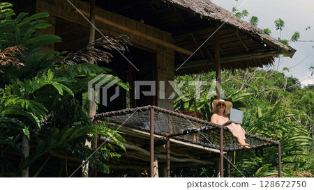 Surrounded by lush greenery, a young woman sits on a terrace of bamboo hut, focusing on her laptop while enjoying the serene atmosphere of a tropical jungle, a perfect mix of work and nature Surrounded by lush greenery, a young woman sits on a terrace of bamboo hut, focusing on her laptop while enjoying the serene atmosphere of a tropical jungle, a perfect mix of work and nature 128672750