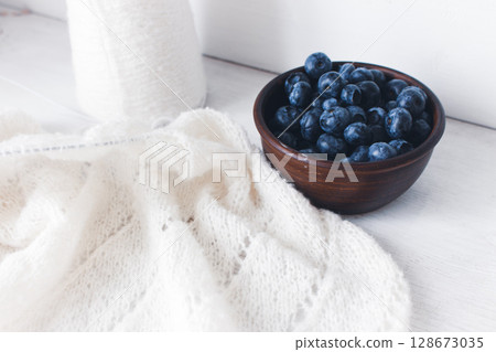 Fresh blueberries in clay bowl and white knitting. Knitted pattern and cotton knitted cardigan. Summer berries and cotton knitted sweater. Summer still life. Organic food and knitting hobby.  128673035