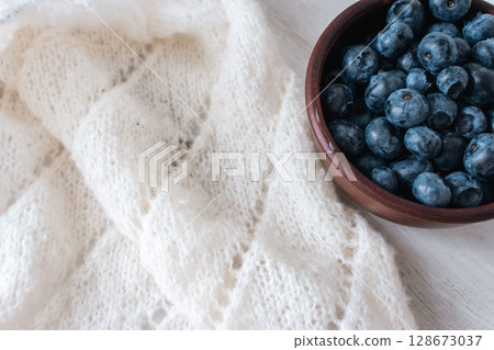 Fresh blueberries in clay bowl and white knitting. Knitted pattern and cotton knitted cardigan. Summer berries and cotton knitted sweater. Summer still life. Organic food and knitting hobby.  128673037