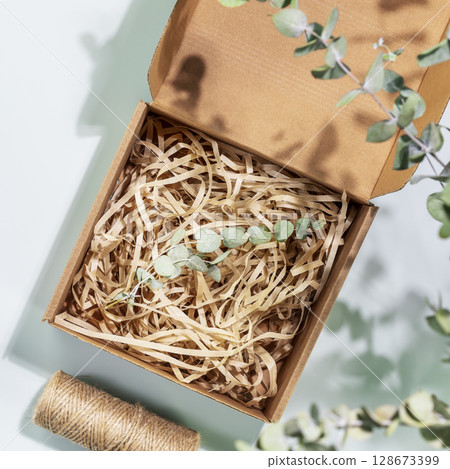 Eco friendly packaging concept. Cardboard box with shredded paper and eucalyptus sprig on blue background with green leaves and shadows. Eco packaging goods from online stores and delivery. Flat lay 128673399