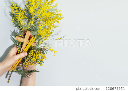 Easter religion card. Womans hands hold wooden crucifixion, mimosa sprig and church candles on blue background. Concept of Holy Week, Palm Sunday, Lent and religion holidays. Flat lay, copy space 128673651
