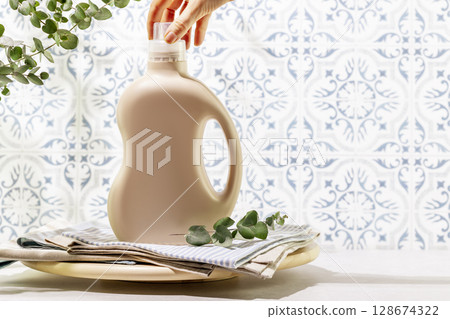 Natural laundry detergent mockup. Sustainable life style with female hande unscrews the lid from bottle with washing gel on textile napkins on wooden podium opposite tile wall. Laundry day. Copy space 128674322
