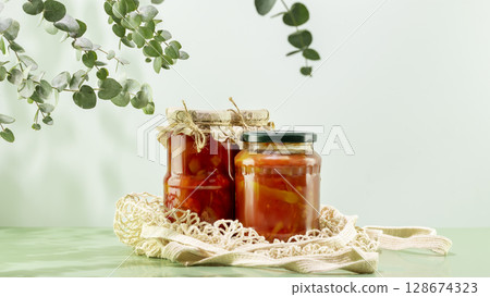 Probiotic foods. Eco friendly still life with pickled or fermented vegetables. Lecho and ajvar in glass jars on textile bag on blue background with leaves and shadows. Home food preserving or canning Probiotic foods. Eco friendly still life with pickled or fermented vegetables. Lecho and ajvar in glass jars on textile bag on blue background with leaves and shadows. Home food preserving or canning 128674323