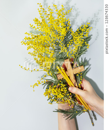 Easter holiday religion card. Womans hands hold wooden crucifixion, mimosa sprig and church candles on blue background. The concept of Holy Week, Palm Sunday, Lent and religion holidays. Flat lay Easter holiday religion card. Womans hands hold wooden crucifixion, mimosa sprig and church candles on blue background. The concept of Holy Week, Palm Sunday, Lent and religion holidays. Flat lay 128674330