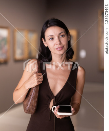 Woman with phone in hands in museum hall, view painting exhibition Woman with phone in hands in museum hall, view painting exhibition 128675468