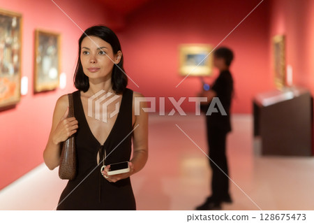 Woman with phone in hands in museum hall, view painting exhibition Woman with phone in hands in museum hall, view painting exhibition 128675473