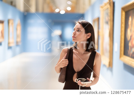 Woman with phone in hands in museum hall, view painting exhibition Woman with phone in hands in museum hall, view painting exhibition 128675477