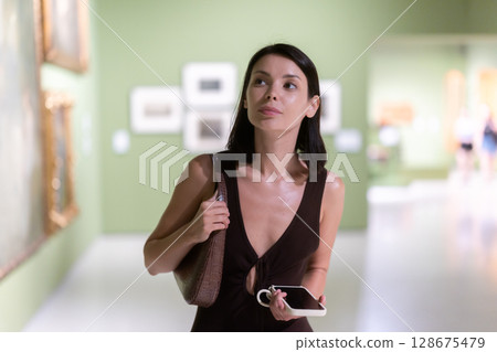 Woman with phone in hands in museum hall, view painting exhibition 128675479