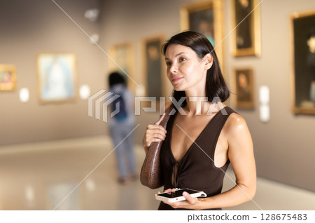Woman with phone in hands in museum hall, view painting exhibition 128675483