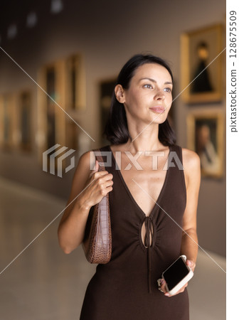 Woman with phone in hands in museum hall, view painting exhibition Woman with phone in hands in museum hall, view painting exhibition 128675509