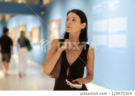 Woman with phone in hands in museum hall, view painting exhibition Woman with phone in hands in museum hall, view painting exhibition 128675564
