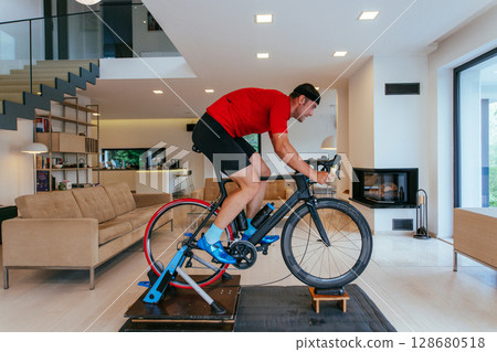 A triathlete riding a triathlon bike on a machine simulation in a modern living room A triathlete riding a triathlon bike on a machine simulation in a modern living room 128680518