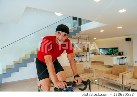 A triathlete riding a triathlon bike on a machine simulation in a modern living room A triathlete riding a triathlon bike on a machine simulation in a modern living room 128680558