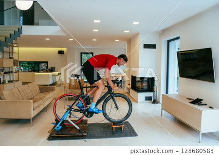 A triathlete riding a triathlon bike on a machine simulation in a modern living room A triathlete riding a triathlon bike on a machine simulation in a modern living room 128680583