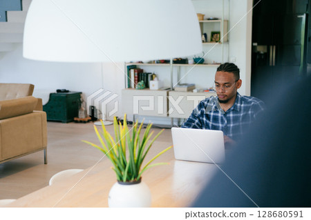 African American influencer in glasses sitting at a table in a modern living room, using a laptop for business video chat, conversation with friends and entertainment. 128680591
