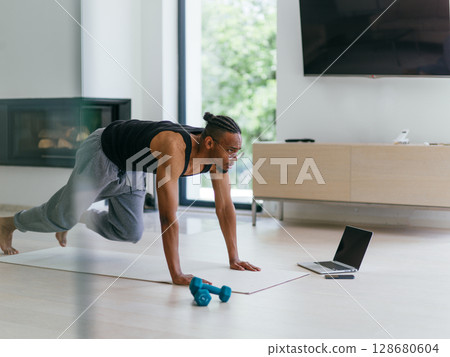 Training At Home. Sporty man doing the training while watching an online tutorial on a laptop, exercising in the living room, free space. 128680604