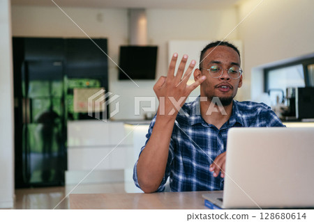 African American influencer in glasses sitting at a table in a modern living room, using a laptop for business video chat, conversation with friends and entertainment. 128680614