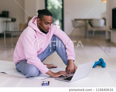 African American man in glasses sitting at a table in a modern living room, using a laptop for business video chat, conversation with friends and entertainment. African American man in glasses sitting at a table in a modern living room, using a laptop for business video chat, conversation with friends and entertainment. 128680616