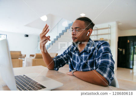 African American influencer in glasses sitting at a table in a modern living room, using a laptop for business video chat, conversation with friends and entertainment. 128680618