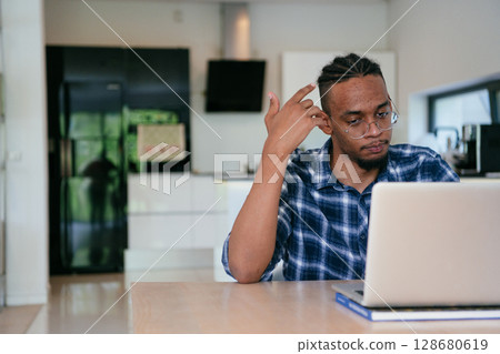 African American influencer in glasses sitting at a table in a modern living room, using a laptop for business video chat, conversation with friends and entertainment. African American influencer in glasses sitting at a table in a modern living room, using a laptop for business video chat, conversation with friends and entertainment. 128680619