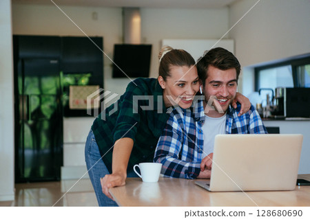 A young married couple is talking to parents, family and friends on a video call via a laptop while sitting in the living room of their modern house. 128680690