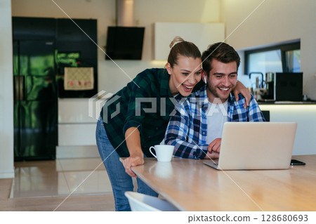 A young married couple is talking to parents, family and friends on a video call via a laptop while sitting in the living room of their modern house. A young married couple is talking to parents, family and friends on a video call via a laptop while sitting in the living room of their modern house. 128680693