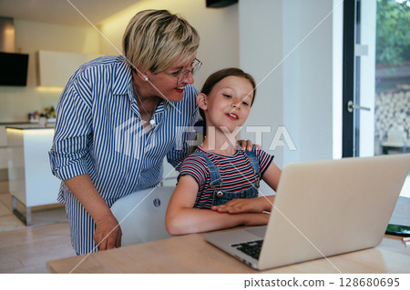 Mother with her daughter talking on laptop with family and friends while sitting in modern living room of big house. Mother with her daughter talking on laptop with family and friends while sitting in modern living room of big house. 128680695