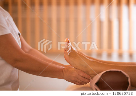 Foot Massage Therapy in a Relaxing Spa Setting. A professional massage therapist in a white uniform provides a soothing foot massage to a client in a calm and serene spa environment Foot Massage Therapy in a Relaxing Spa Setting. A professional massage therapist in a white uniform provides a soothing foot massage to a client in a calm and serene spa environment 128680738