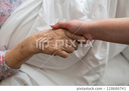 A young womans hands gently hold the hands of an elderly female patient in a hospital bed, symbolizing care, support, and compassion. 128680741