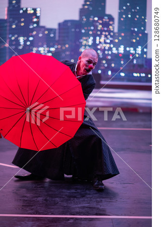 Mysterious Clown Performer with Red Umbrella in Urban Nightscape. Mysterious Clown Performer with Red Umbrella in Urban Nightscape. 128680749