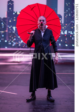 Mysterious Clown Performer with Red Umbrella in Urban Nightscape. Mysterious Clown Performer with Red Umbrella in Urban Nightscape. 128680751