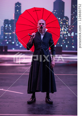 Mysterious Clown Performer with Red Umbrella in Urban Nightscape. Mysterious Clown Performer with Red Umbrella in Urban Nightscape. 128680752