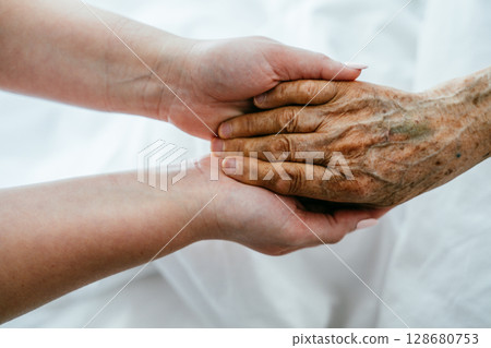 A young womans hands gently hold the hands of an elderly female patient in a hospital bed, symbolizing care, support, and compassion. 128680753