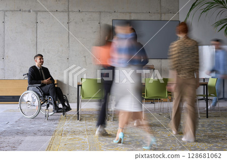 A diverse business team, working urgently around a conference table, collaborates on a meeting while their director, seated in a wheelchair, waits for their input 128681062