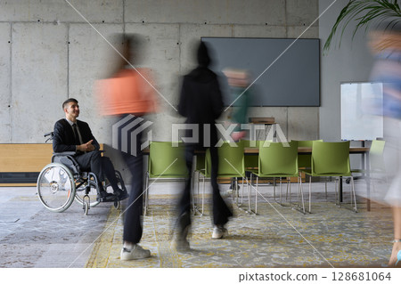A diverse business team, working urgently around a conference table, collaborates on a meeting while their director, seated in a wheelchair, waits for their input 128681064