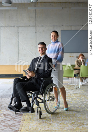 In a professional office setting, a businesswoman helping her director, who is sitting in a wheelchair and using a tablet, while their colleagues collaborate in the background. In a professional office setting, a businesswoman helping her director, who is sitting in a wheelchair and using a tablet, while their colleagues collaborate in the background. 128681077