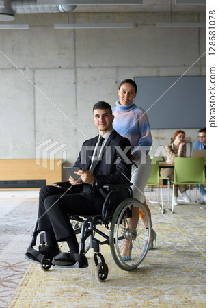 In a professional office setting, a businesswoman helping her director, who is sitting in a wheelchair and using a tablet, while their colleagues collaborate in the background. In a professional office setting, a businesswoman helping her director, who is sitting in a wheelchair and using a tablet, while their colleagues collaborate in the background. 128681078