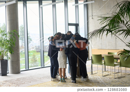 A diverse group of professionals standing in a supportive circle, embracing each other in a modern office space filled with natural light.  128681080