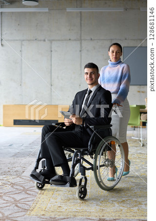 In a professional office setting, a businesswoman helping her director, who is sitting in a wheelchair and using a tablet, while their colleagues collaborate in the background. 128681146