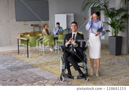 In a professional office setting, a businesswoman helping her director, who is sitting in a wheelchair and using a tablet, while their colleagues collaborate in the background. In a professional office setting, a businesswoman helping her director, who is sitting in a wheelchair and using a tablet, while their colleagues collaborate in the background. 128681153