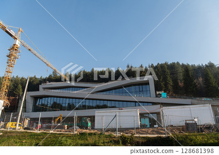 The exterior of a business building under construction is surrounded by trucks, cranes, and various construction materials, creating a bustling scene of an expansive industrial worksite in progress 128681398