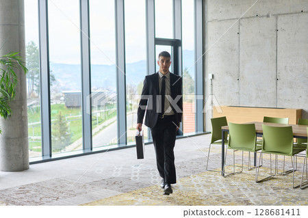 A determined businessman in a black suit confidently walks through the office, embodying professionalism and focus as he heads towards new challenges A determined businessman in a black suit confidently walks through the office, embodying professionalism and focus as he heads towards new challenges 128681411