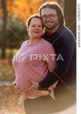 A man lovingly embraces his pregnant wife in the park, sharing a peaceful moment of warmth and affection  128681471