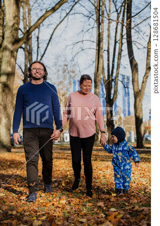 A loving couple strolls through a sunlit park with their young son, surrounded by the vibrant colors of autumn, enjoying a joyful and peaceful family moment together. 128681504