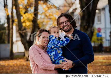 On a sunny autumn day in the park, a mother and father lovingly embrace their young son, sharing moments of laughter and joy as they play together 128681527