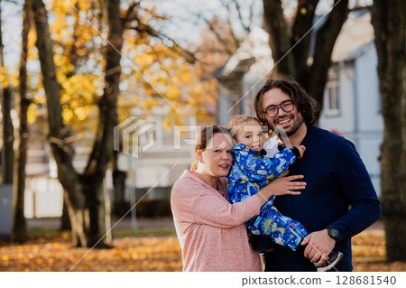 On a sunny autumn day in the park, a mother and father lovingly embrace their young son, sharing moments of laughter and joy as they play together 128681540